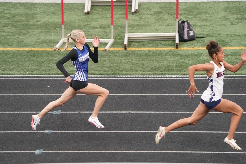 Two female track athletes sprinting on a track during a race, with one athlete in a black and blue uniform and the other in a white and blue uniform.