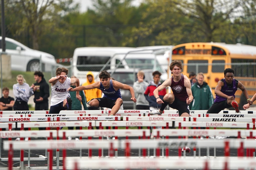 High school athletes competing in a hurdle race on an outdoor track, with spectators in the background.