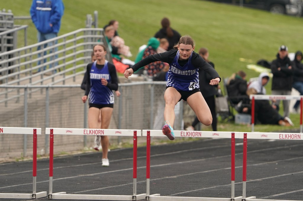 A female athlete competing in a hurdle race, leaping over a hurdle while another runner follows behind.