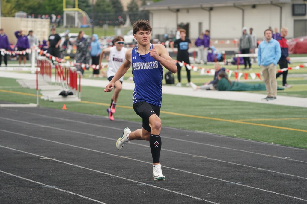 A young male athlete sprinting on a track during a competition, wearing a blue sports uniform with the name 'Bennington' printed on it.