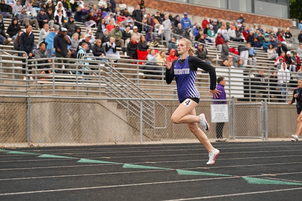 A female athlete sprinting on a track during a race, wearing a purple and black uniform with the number 4. Spectators are visible in the stands, dressed in various clothing for outdoor weather.