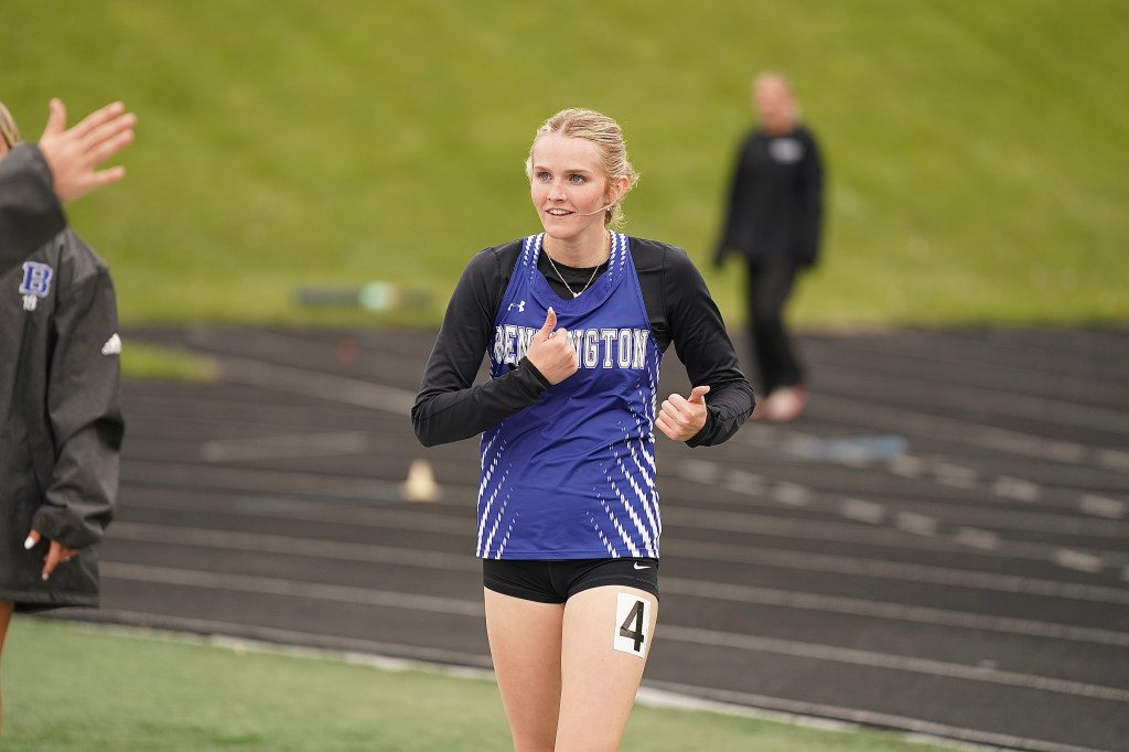 A female athlete in a blue and black running jersey smiles and gives a thumbs up while on a track field.