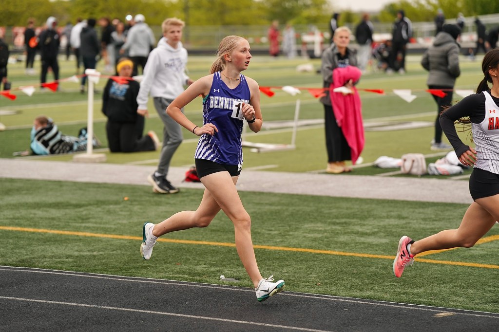 A female athlete wearing a blue and white jersey with the number 11 is running on a track during a competition, with other athletes and spectators in the background.