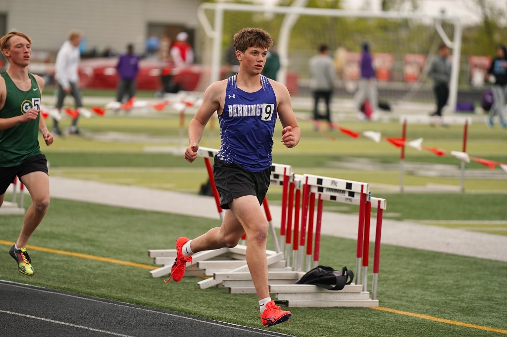 A male athlete in a blue tank top with the number 9 is sprinting on a track during a hurdle race, with another runner in a green shirt in the background.