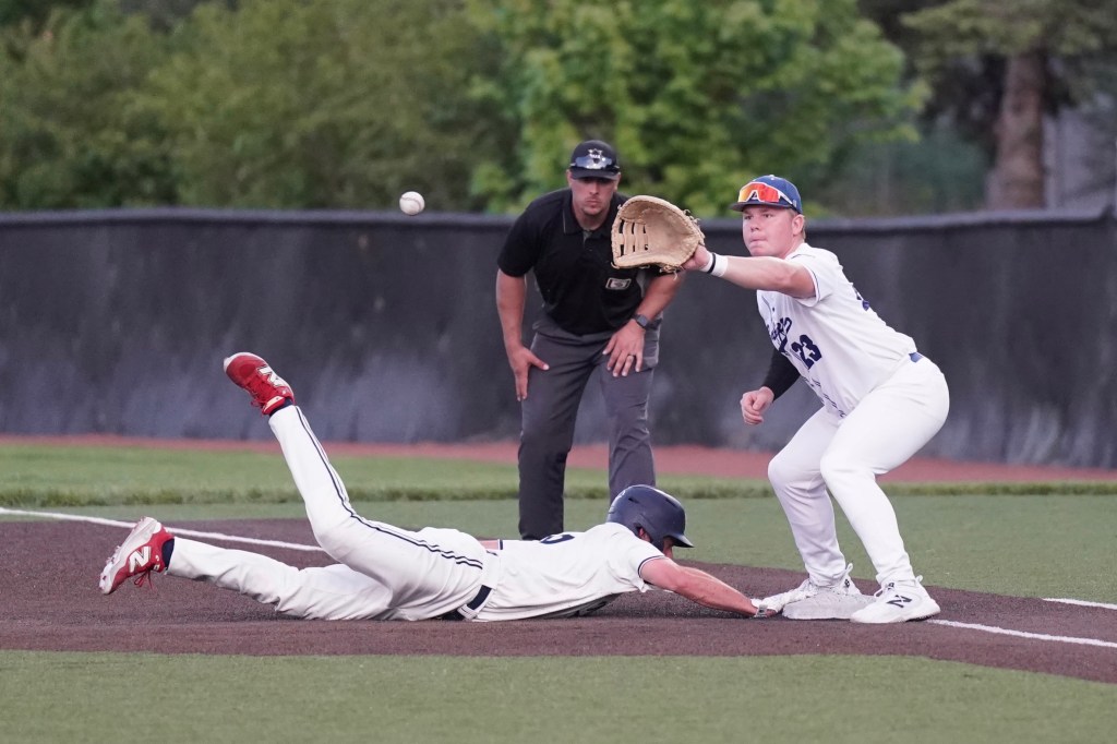 A baseball player slides into base while another player reaches out to catch a thrown ball, with an umpire watching the play from behind.