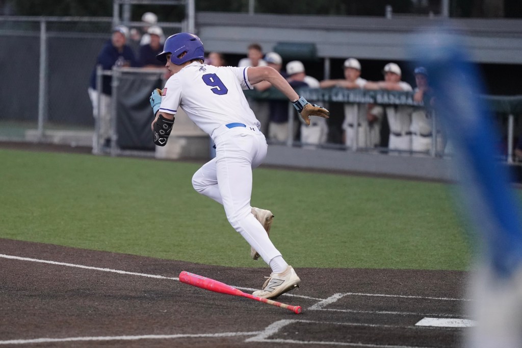 A baseball player in a white uniform with the number 9 is running towards first base, holding a bat, while teammates watch from the dugout.