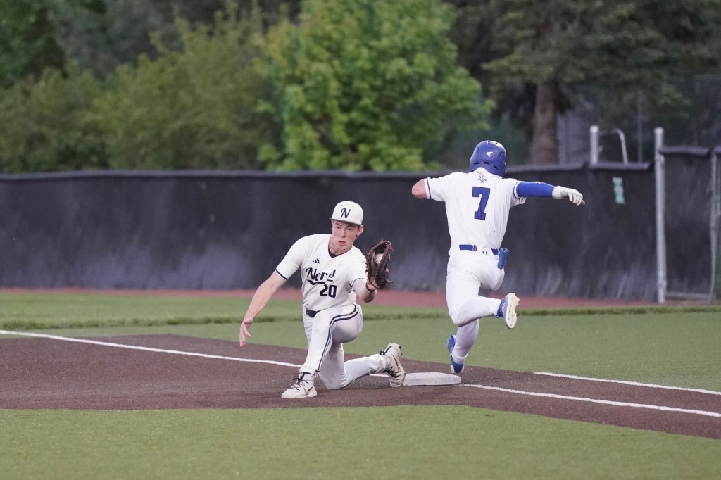A baseball player in a white uniform is attempting to tag out a runner in blue who is sliding towards the base. The scene is set on a well-maintained baseball field with trees in the background.