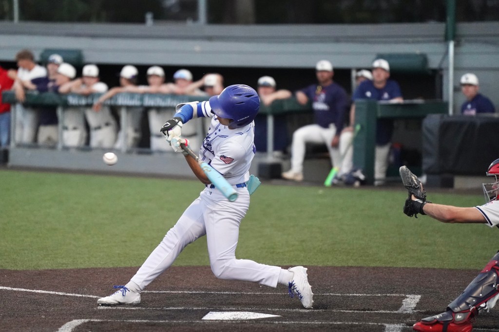 A young baseball player swings a bat at a pitch during a game, with teammates in the background watching from the dugout.