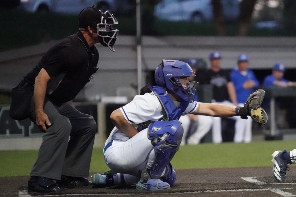 A baseball catcher in blue gear crouches to catch a pitch while the home plate umpire in black stands nearby. The background shows players in team uniforms.