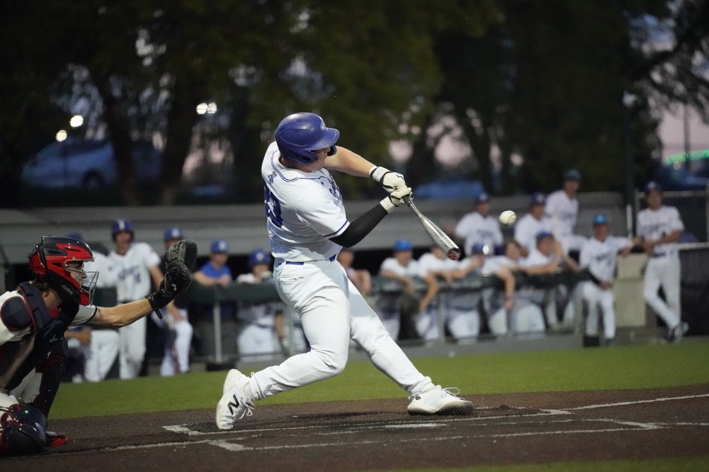 A baseball player in a white uniform swings a bat at a baseball during a game, while the catcher, wearing a red helmet and chest protector, crouches behind home plate.