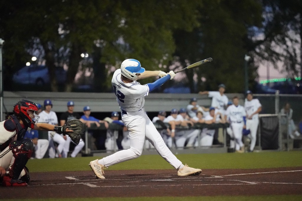 A baseball player swings for a hit during a game, while a catcher crouches behind home plate. The scene captures other players in the dugout cheering in the background.