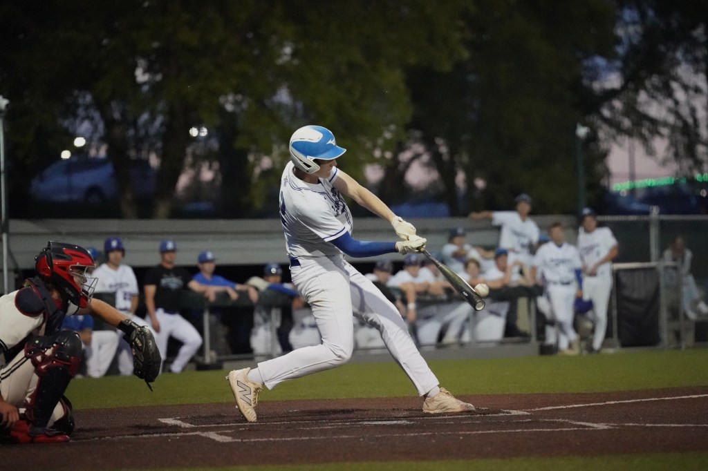 A baseball player swings a bat while a catcher prepares to catch the pitch, with teammates in the background watching the game.