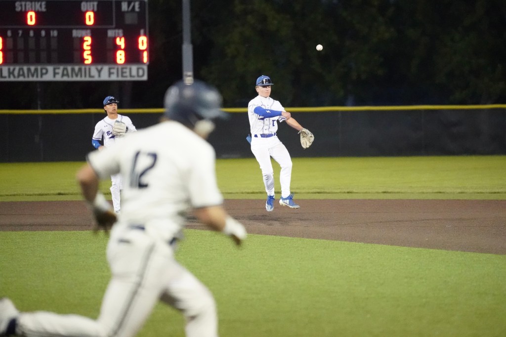 A baseball player in a white uniform is running towards home plate while a teammate in a blue and white uniform is preparing to throw the ball from the infield. Another player stands in the background, ready in position. The scoreboard shows the game status.