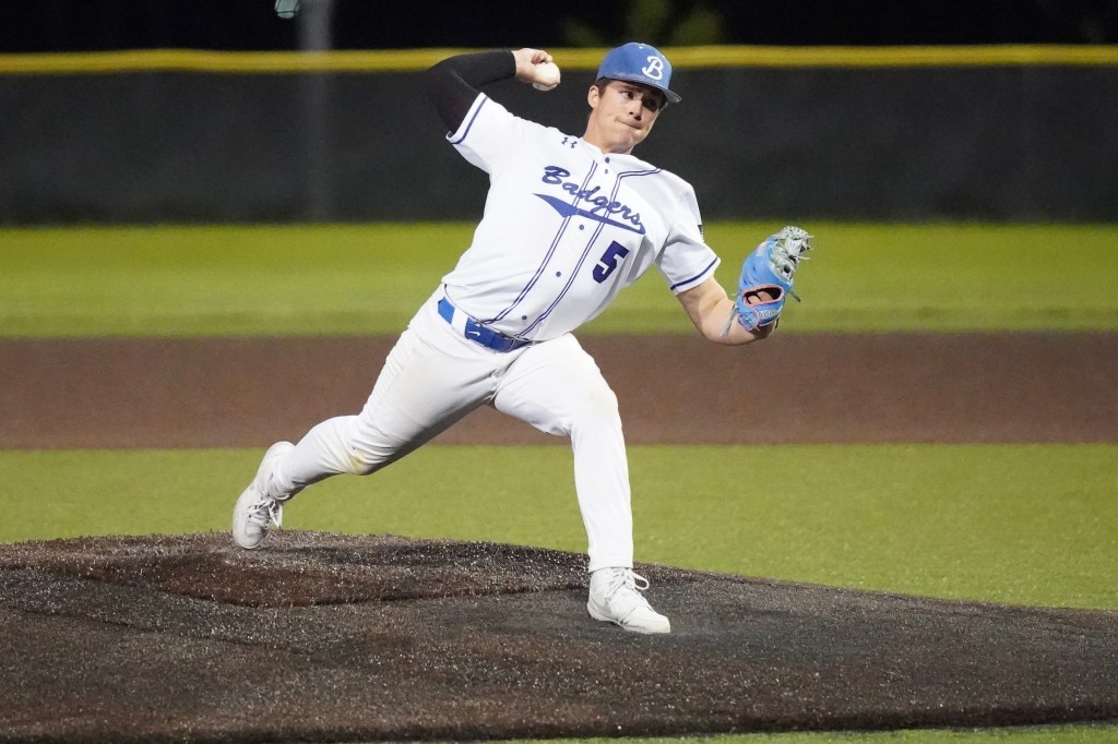 A baseball player wearing a white jersey with blue accents pitches the ball on a baseball mound at night.