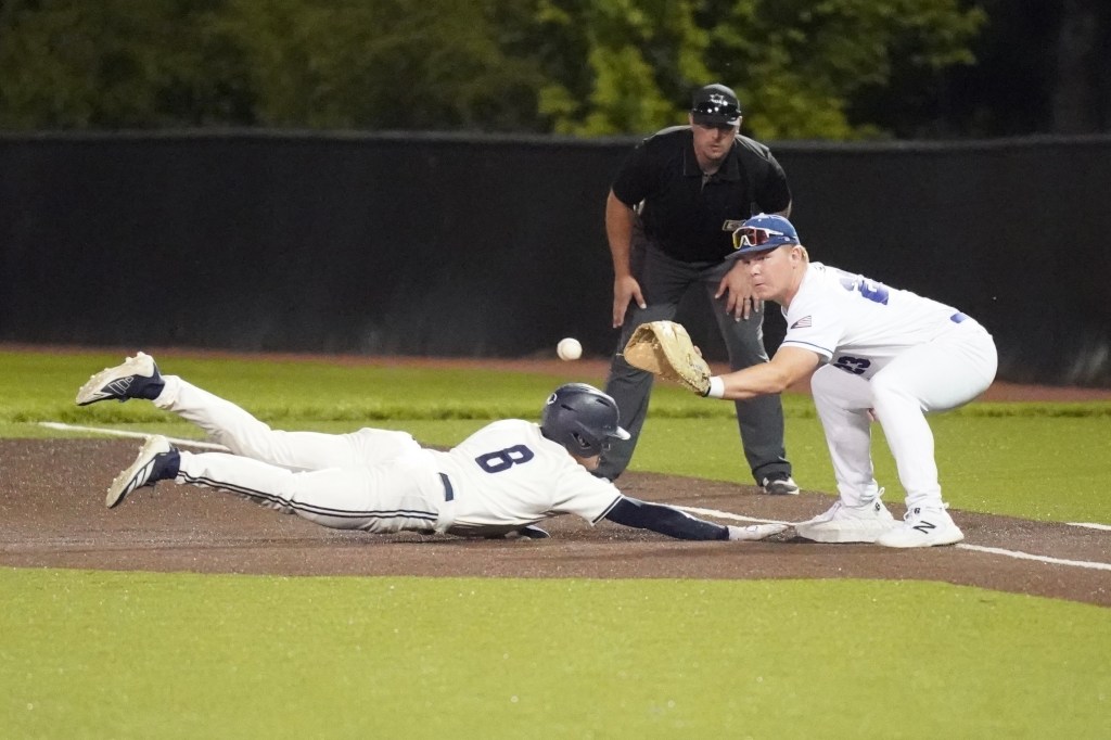 A baseball player slides towards a base while another player prepares to catch the ball, with an umpire observing the play in the background.