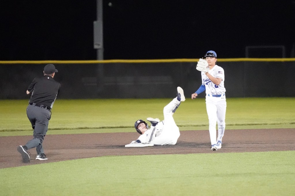 A baseball player slides into second base while another player stands ready to catch the ball, with an umpire observing the play.