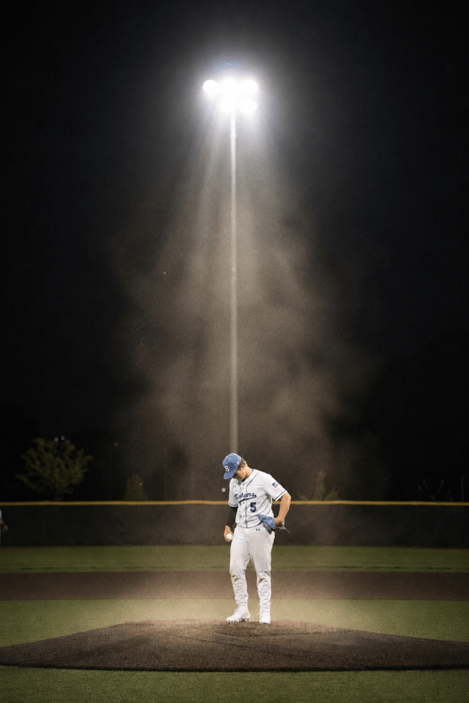 A baseball pitcher stands on the mound at night, illuminated by a bright floodlight, with dust swirling around him.