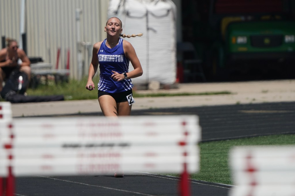 A female athlete in a blue tank top with 'Bennington' written on it, running on a track near hurdles on a sunny day.