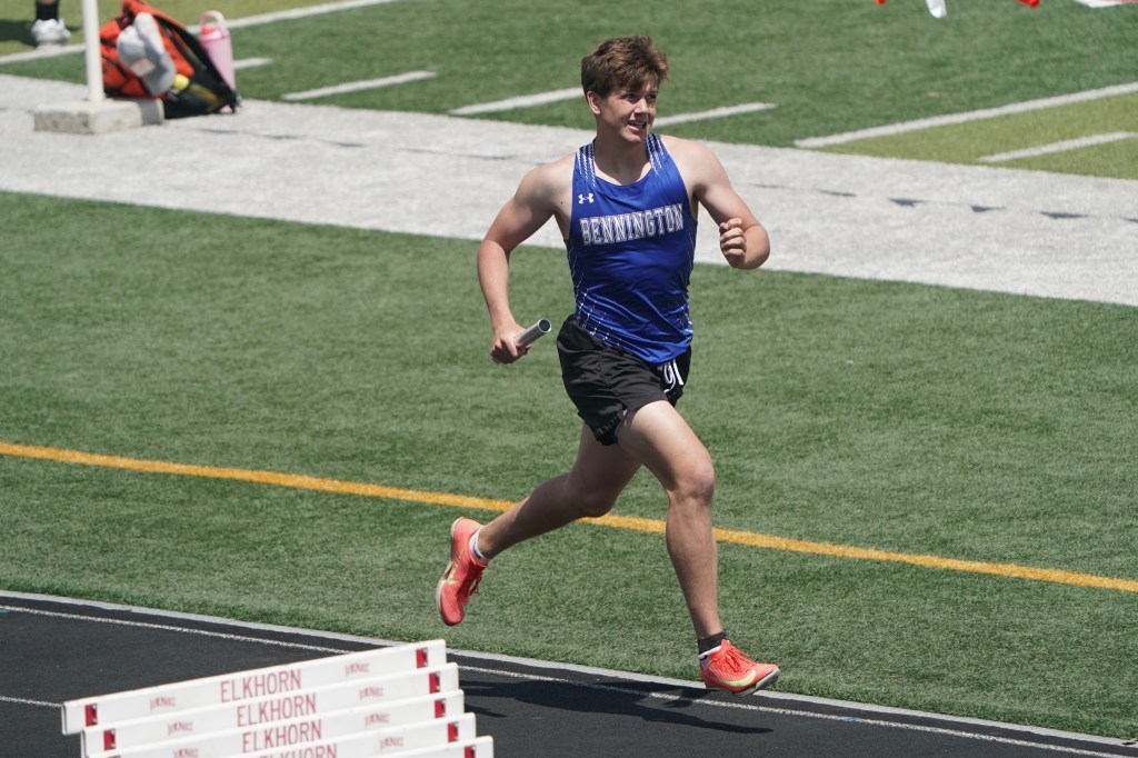 A young male athlete in a blue sports jersey runs on a track during a relay race, holding a baton in his right hand.