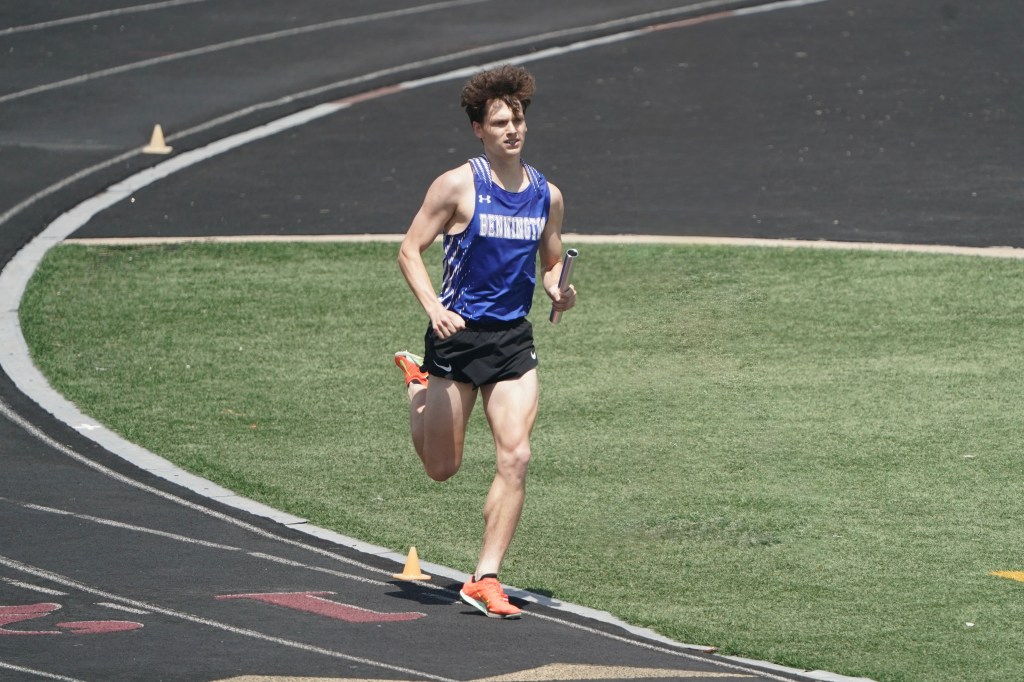 A young male runner in a blue athletic shirt and black shorts competes on a track, holding a relay baton.