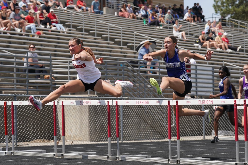 Two female athletes racing in a hurdle event on a track, with spectators in the background.