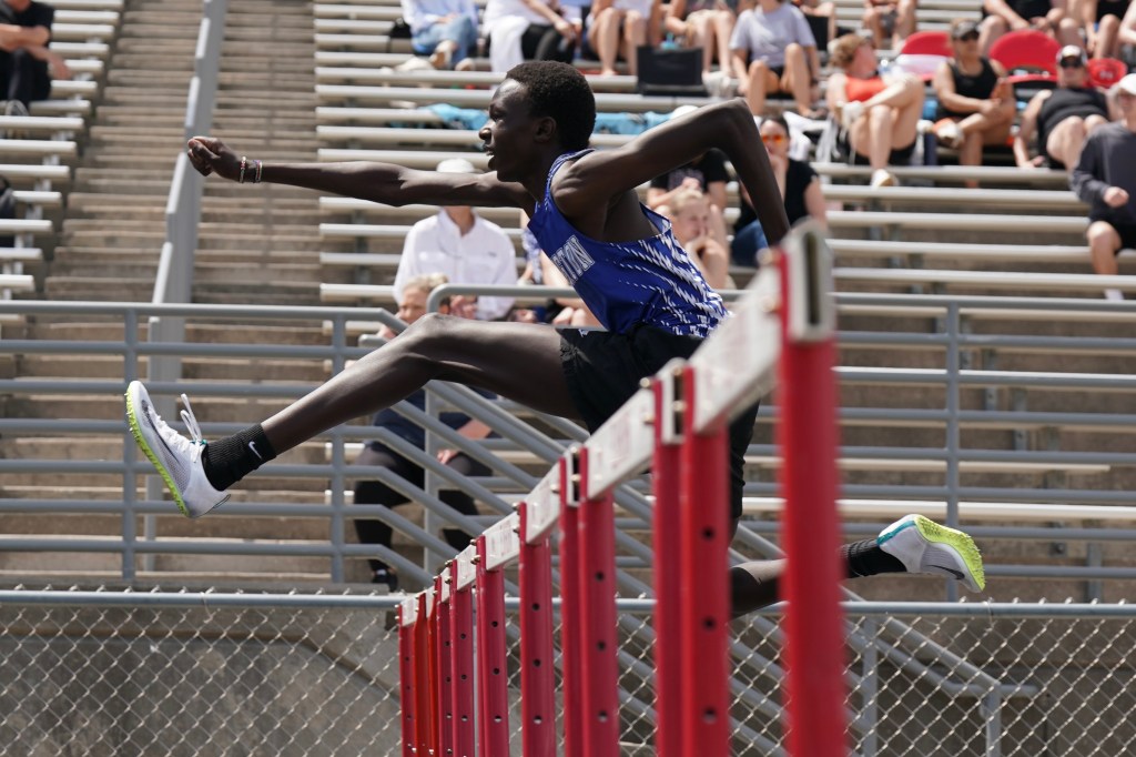 A male athlete jumping over a hurdle during a track and field event, with a crowd in the background.