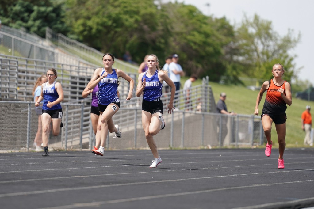 A group of female track athletes competing in a race on a sunny day, with runners in blue uniforms leading the pack.