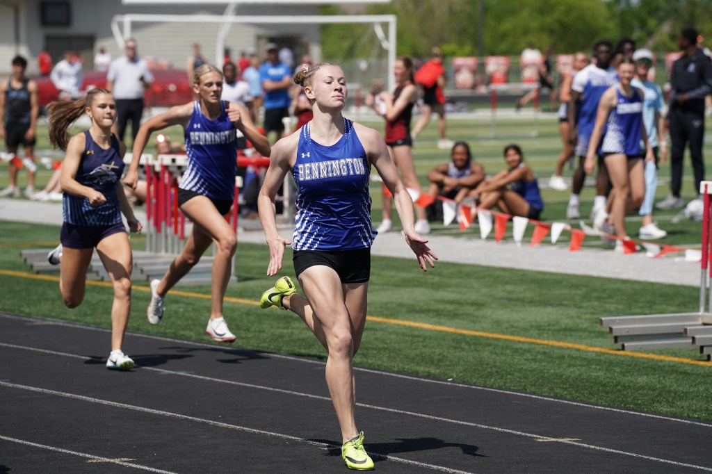 A group of female runners competing in a track event, with one athlete in the foreground from Bennington wearing a blue jersey, focused on finishing the race.