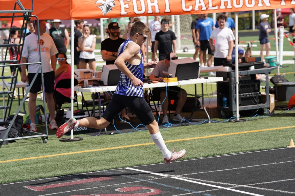 A high school athlete running on a track during a competition, with a group of spectators and equipment in the background.
