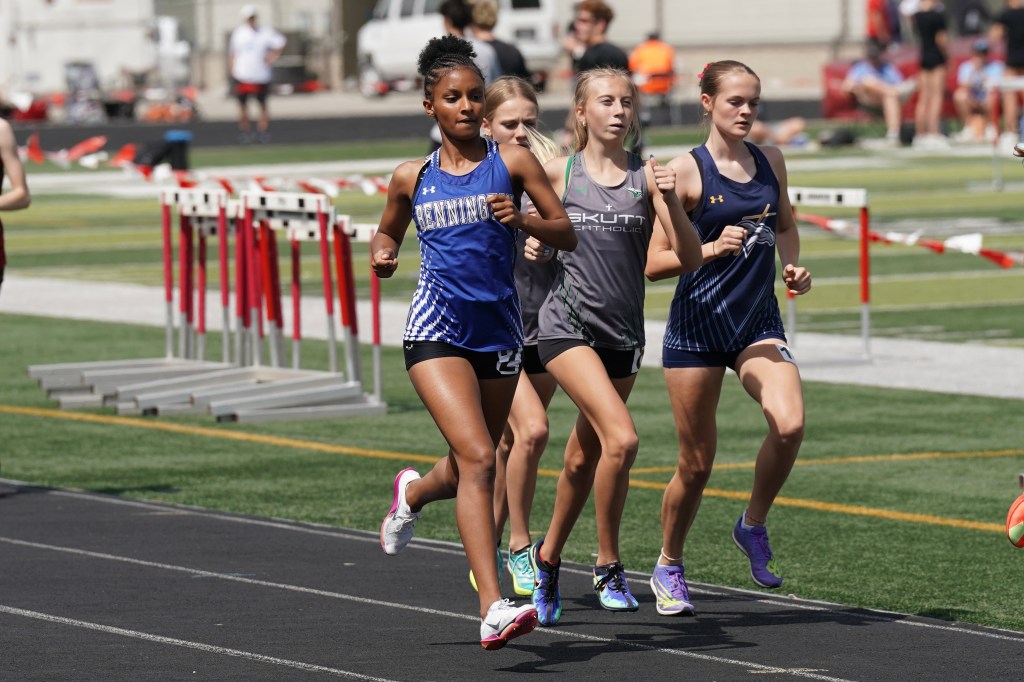 Four female track athletes running on an outdoor track during a competition, with hurdles in the background.