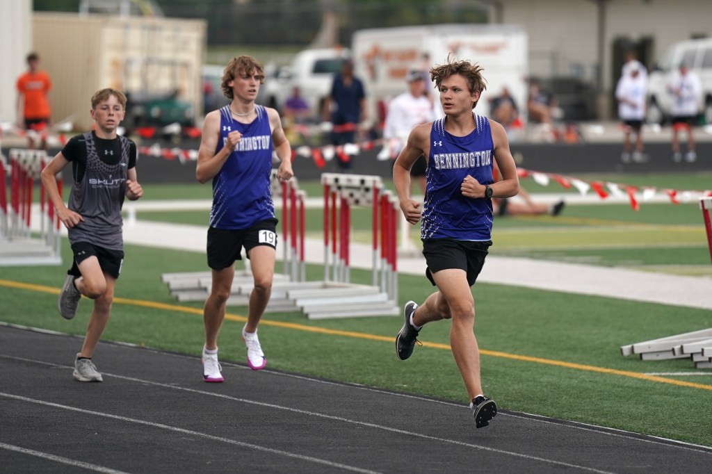 Three male athletes competing in a track and field race, running on a track lined with hurdles. The athlete in the foreground is wearing a blue jersey and black shorts, while the other two athletes wear dark jerseys. The setting appears to be an outdoor athletic event.