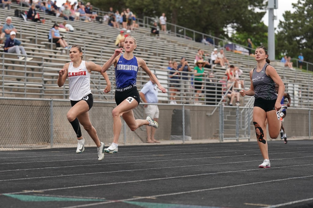 Three female track athletes sprinting on a running track during a race, with spectators in the background.