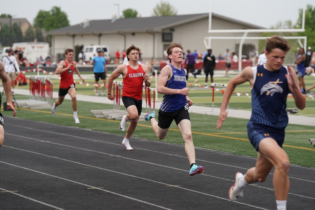 High school track athletes competing in a sprint race on a black track, with several runners wearing different uniforms and approaching hurdles in the background.