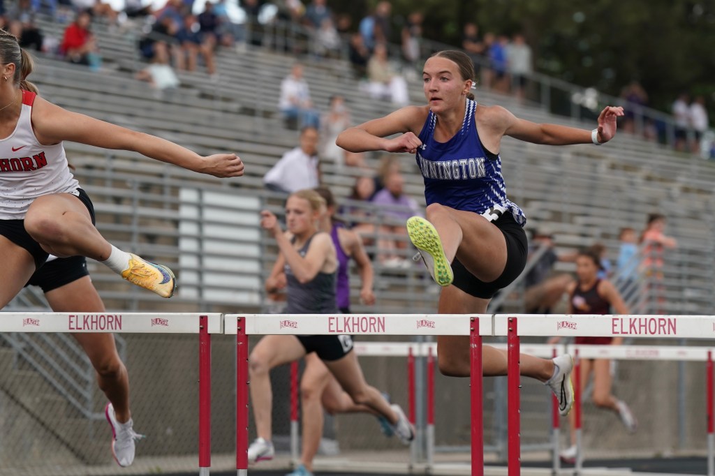 A female athlete competing in a hurdle race, jumping over a barrier while other runners are visible in the background.