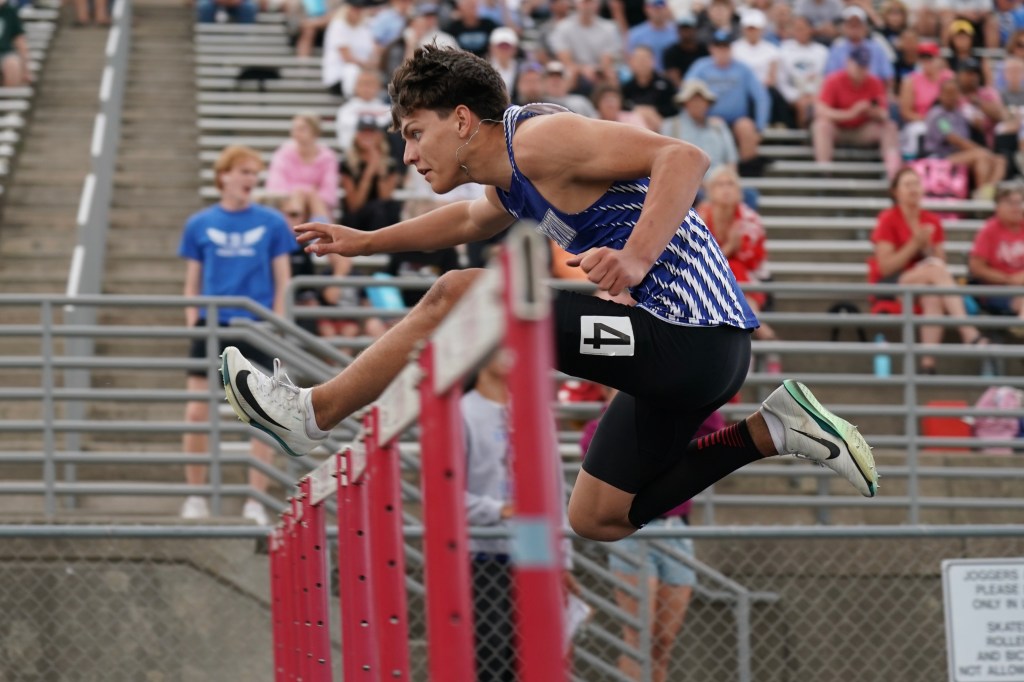 A high school athlete competing in a hurdle race, jumping over a red hurdle with a focused expression, while spectators watch from the stands.