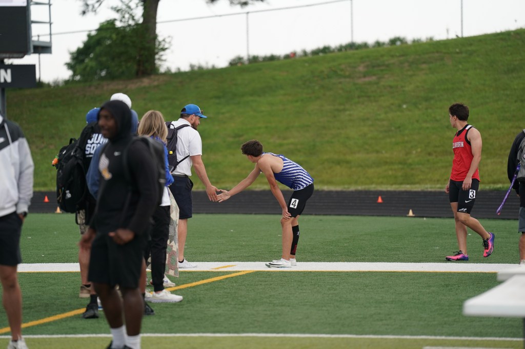 Two athletes preparing for a race on a sports field, with one athlete in a striped jersey leaning forward and another reaching out to shake hands. Several spectators and participants are visible in the background.