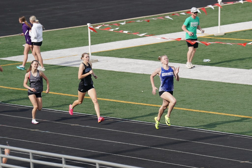 A group of female track runners competing on a track during a race, with one runner in a blue jersey leading the way.