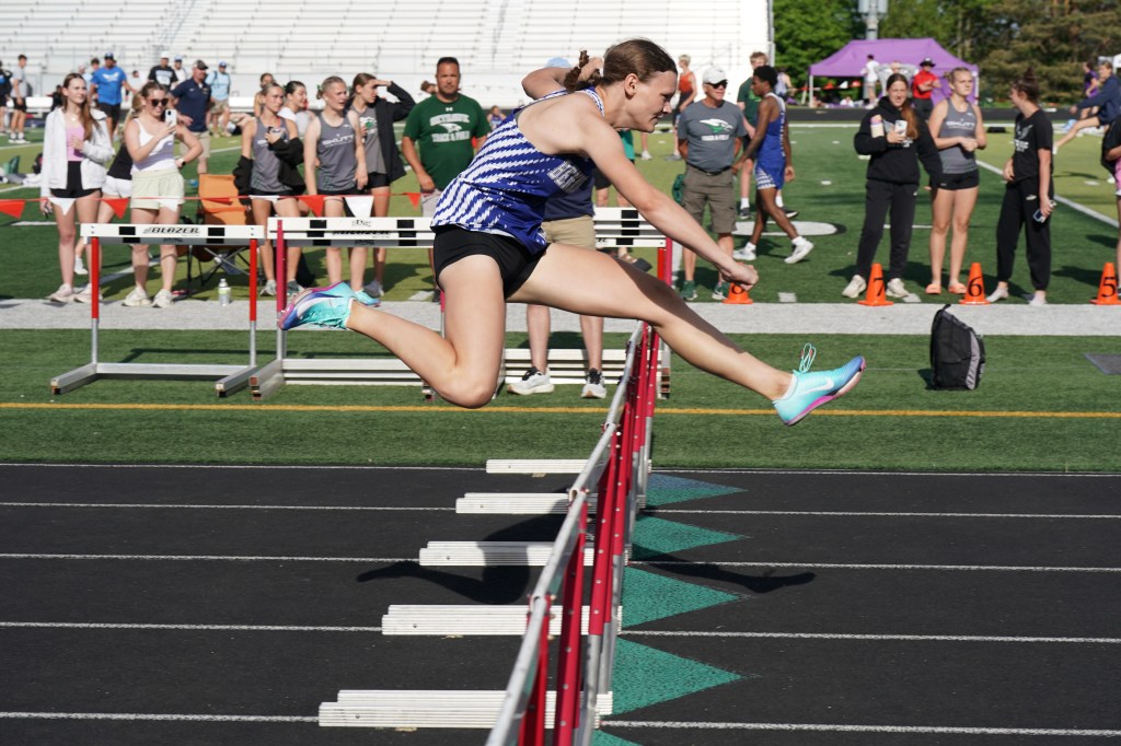 A female athlete competes in a hurdle race, leaping over a red hurdle on a track, with spectators in the background.