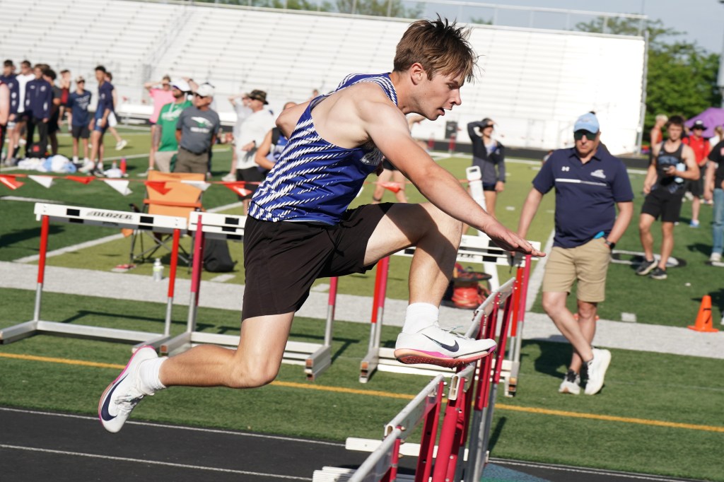 A male athlete competing in a hurdle race, leaping over a hurdle on a track field, with spectators in the background.