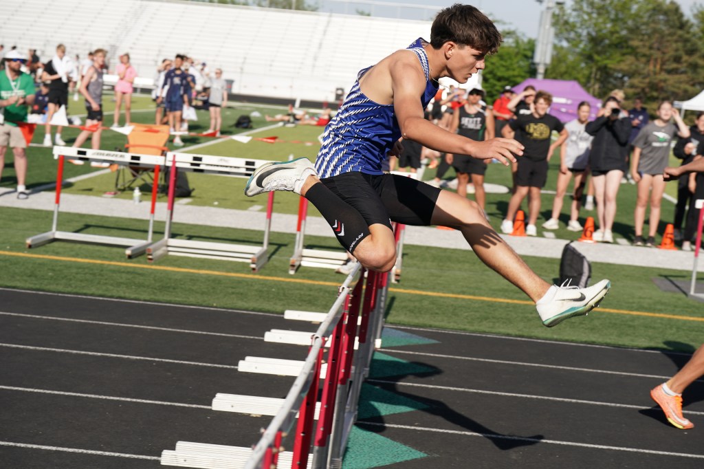 A male athlete in a blue and black uniform hurdles over a red barrier during a track and field event, with spectators in the background.