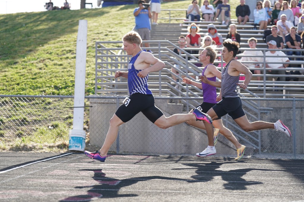 Three male athletes sprinting on a track during a race, with spectators in the background.
