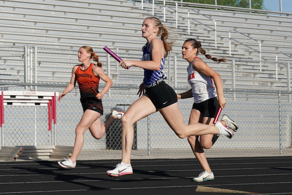 Three female athletes sprinting on a track during a relay race, one holding a baton and wearing a blue uniform.