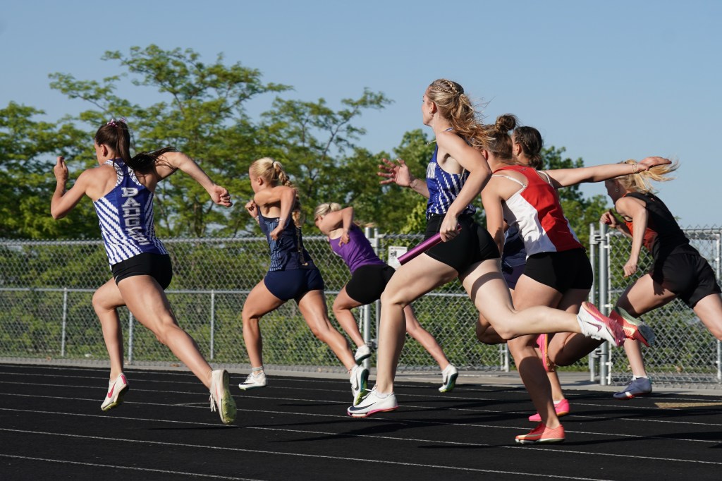 A group of female athletes competing in a relay race on an outdoor track, captured mid-sprint with a clear blue sky in the background.