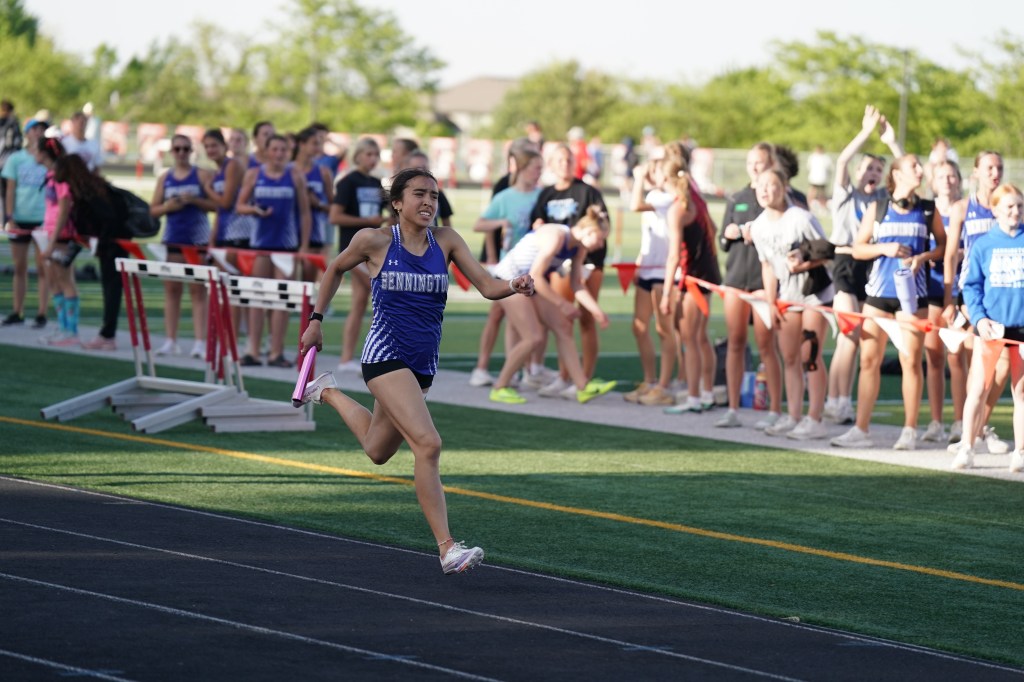 A high school female athlete from Bennington races on a track, holding a relay baton, while teammates and spectators cheer in the background.