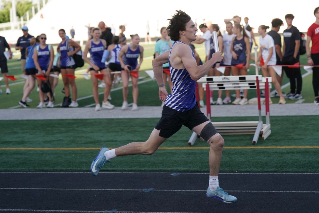 A male athlete in a blue and white uniform sprinting on a track during a relay race, with a group of spectators and other athletes in the background.