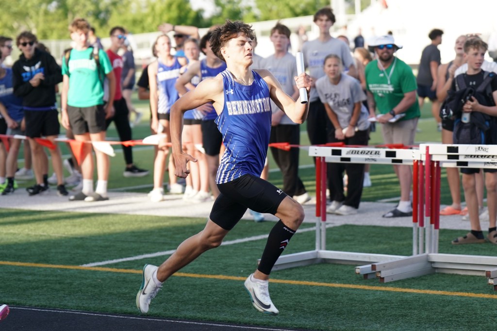 A male athlete in a blue sports uniform running during a track event, holding a relay baton, with spectators in the background.