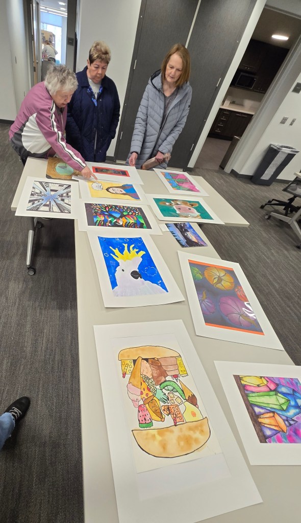 Three women engaging with a display of colorful art pieces on a table in a well-lit room.