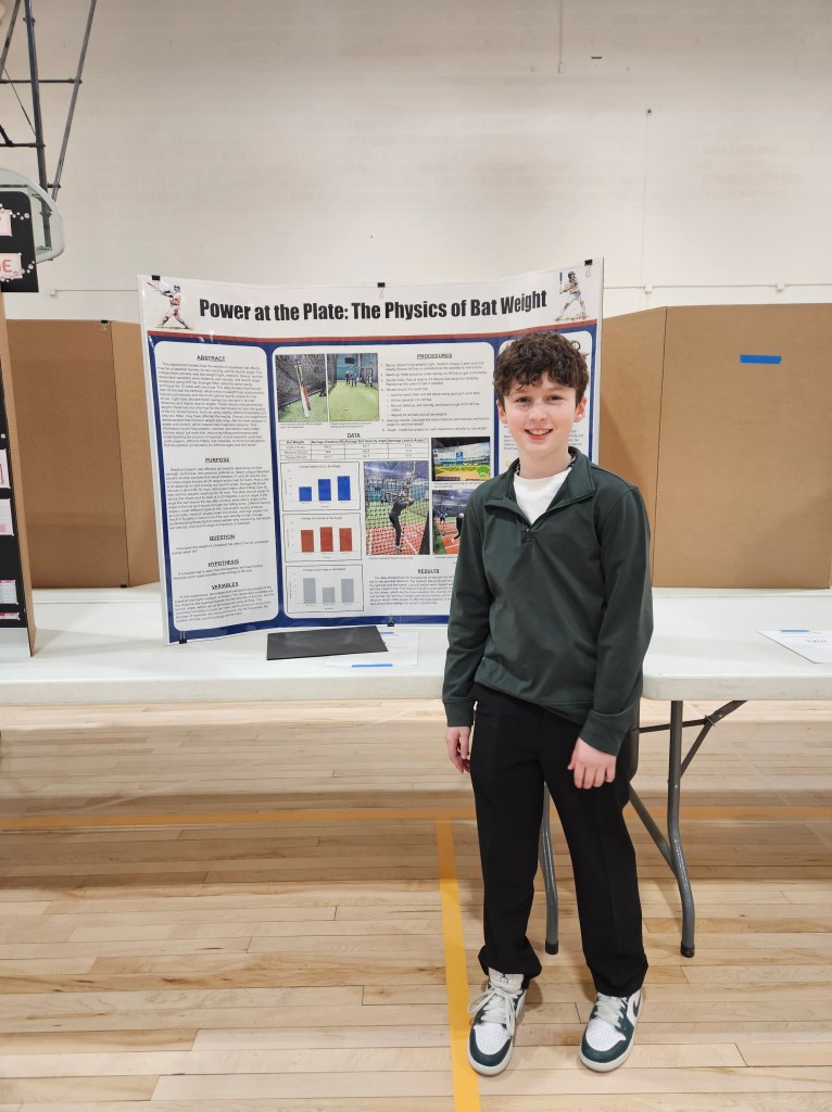 A young boy stands beside a science fair project titled 'Power at the Plate: The Physics of Bat Weight,' which features research findings and graphics related to the topic.