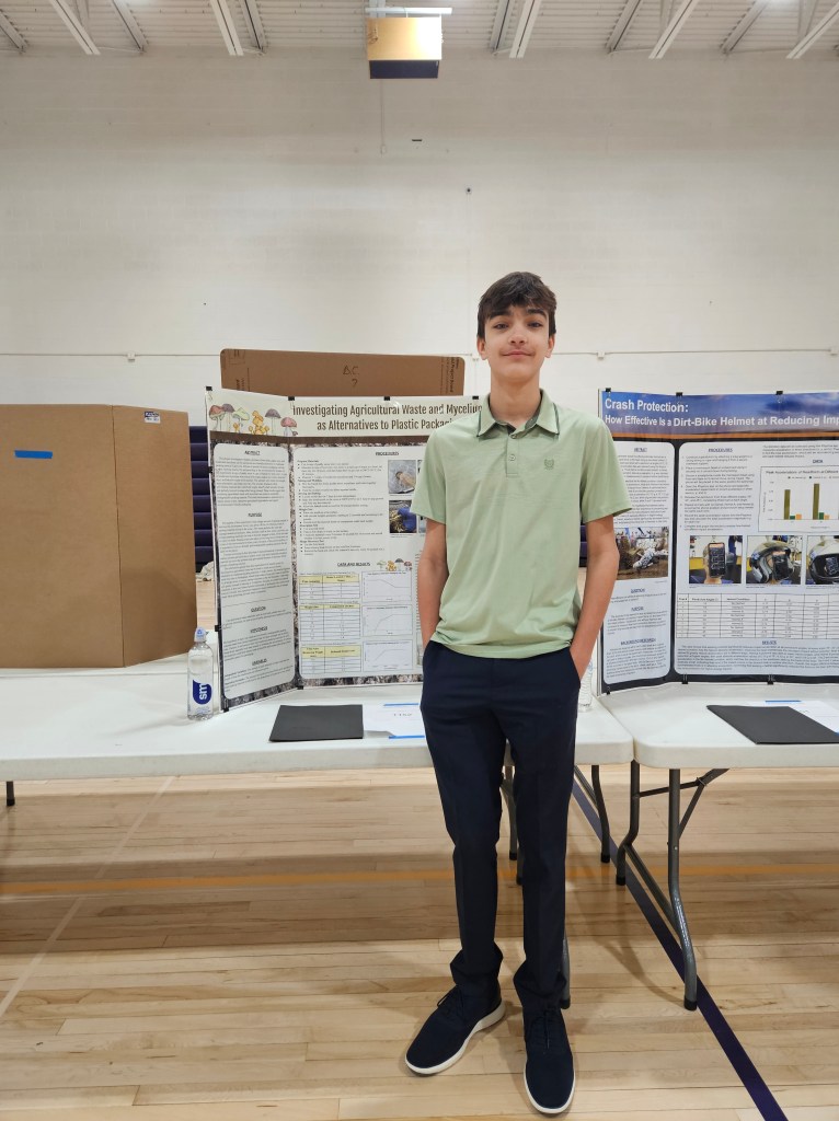 A young male student standing in front of science project display boards in a gymnasium, wearing a light green polo shirt and dark pants.