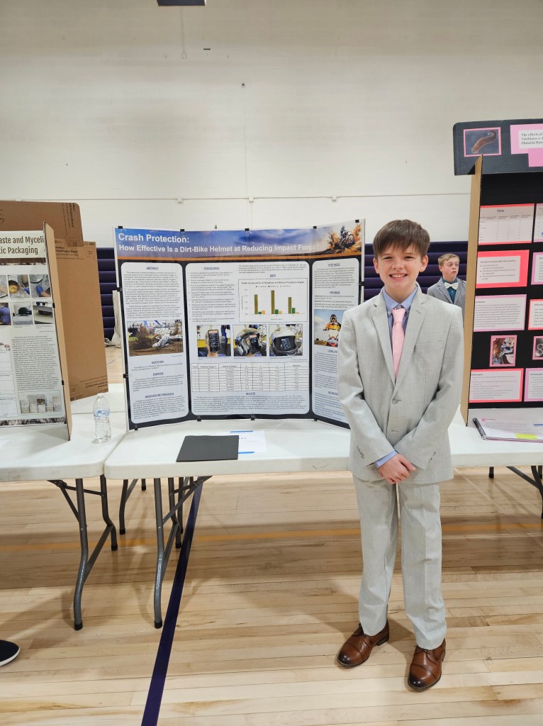 A young boy in a light gray suit and pink tie stands beside a science project display titled 'Crash Protection: How Effective Is a Dirt-Bike Helmet at Reducing Impact Force.' The background shows a gymnasium setting with other project boards.